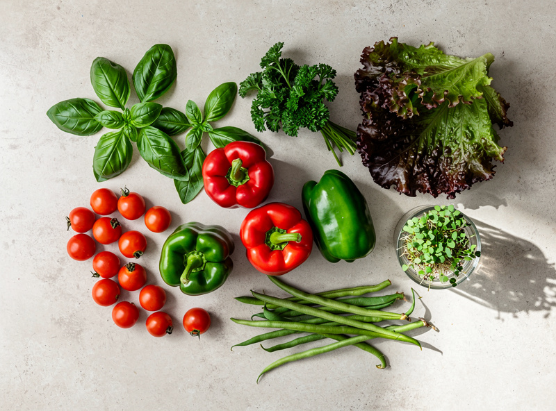 A bountiful overhead view of harvested produce — cherry tomatoes, peppers, basil, parsley, lettuce, beans, and microgreens on a city kitchen counter